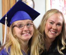 A female student wearing graduation gown, standing next to his mother, and both are smiling